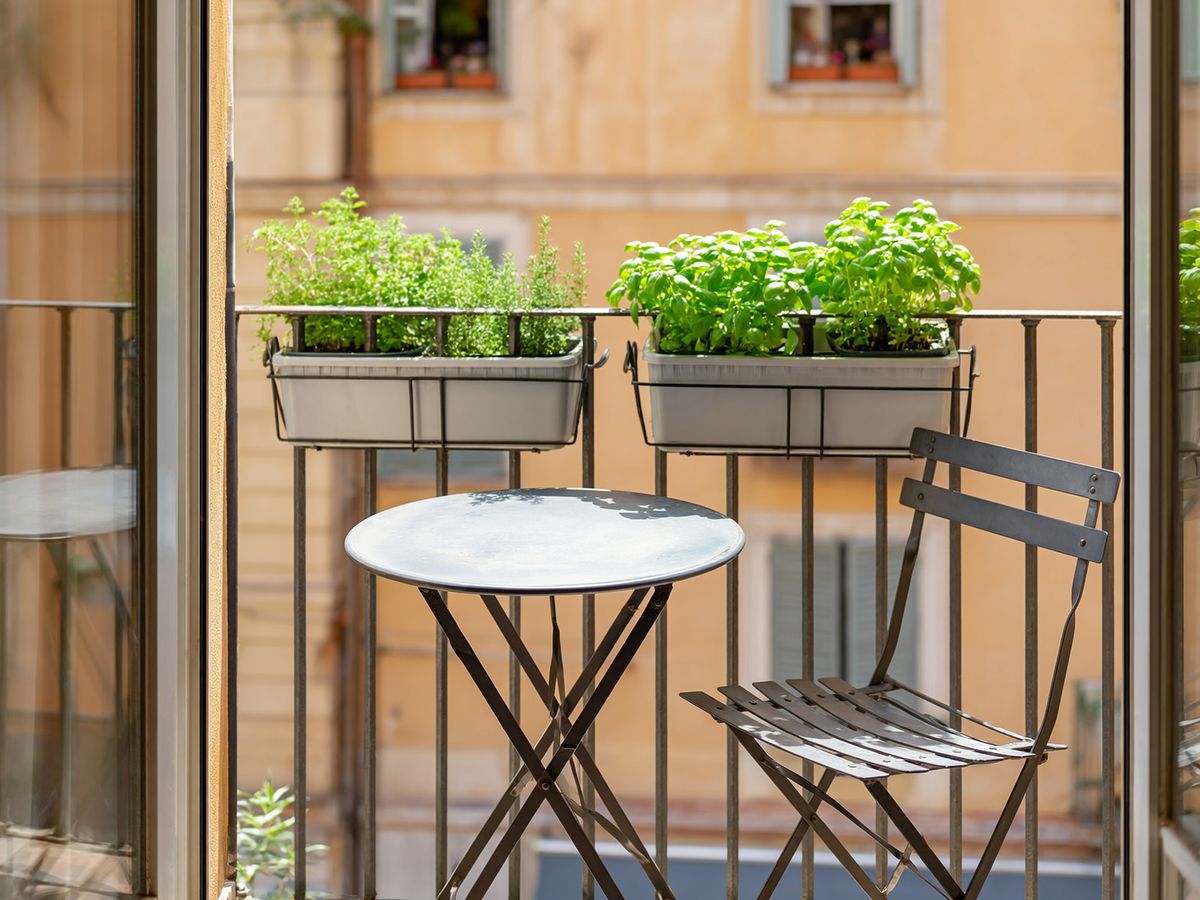 Un piccolo balcone cittadino ben organizzato, con un tavolino in metallo da bistrot, una sedia pieghevole e vasi rettangolari sospesi alla ringhiera riempiti di erbe aromatiche verdi e rigogliose, senza ostacolare il passaggio.